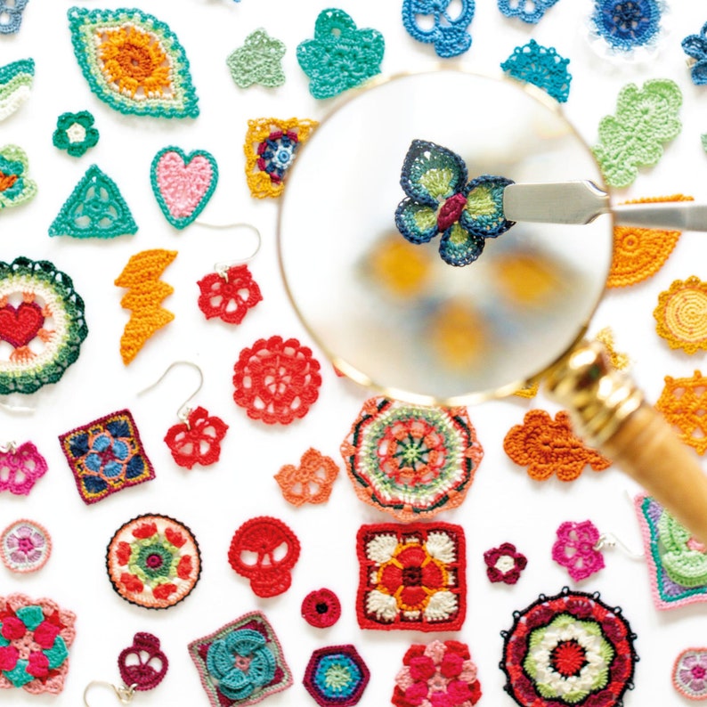 K&ouml;nnte beinhalten: Eine Sammlung bunter geh&auml;kelter Ornamente, darunter Blumen, Herzen, Schmetterlinge und geometrische Formen. Die Ornamente sind auf einem wei&szlig;en Hintergrund angeordnet, wobei eine Lupe auf ein blaues und gr&uuml;nes Schmetterlingsornament fokussiert ist.