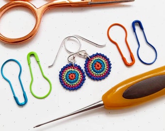 May include: A pair of crocheted earrings with a colourful mandala design. The earrings are blue, green, yellow, orange, and red. The earrings are hanging from silver hooks.
