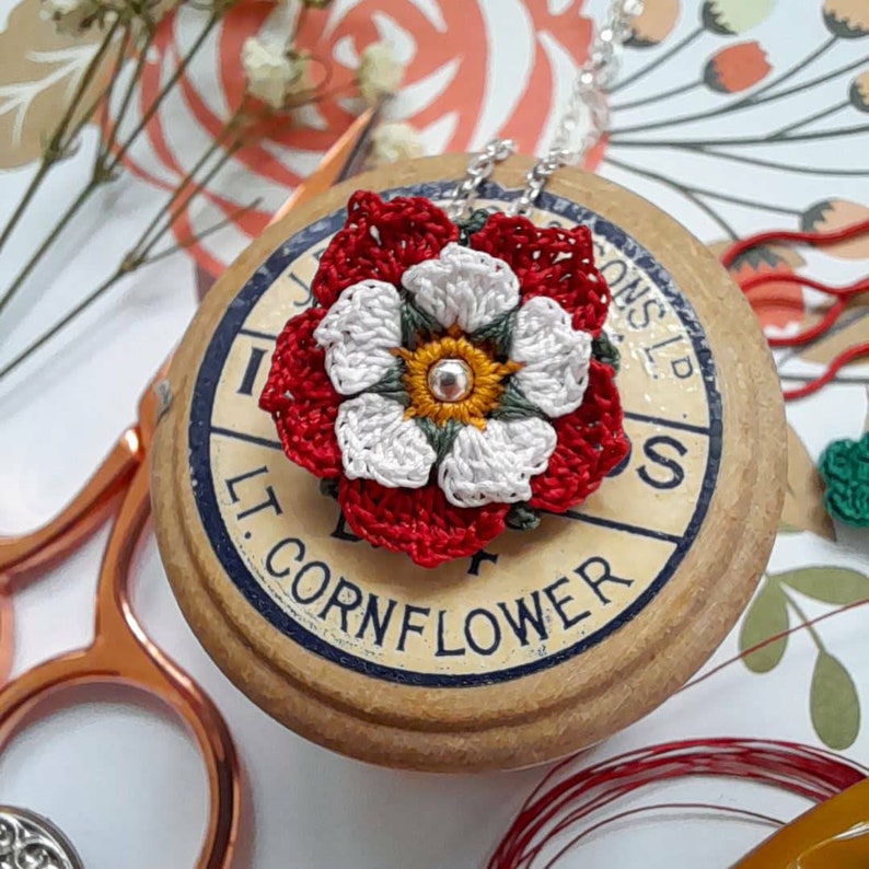 Op de afbeelding: Een witte gehaakte bloem met een rode gehaakte buitenlaag en een gele kern. De bloem is bevestigd aan een zilveren kettingketting. De ketting ligt op een houten spoel met de tekst "LT. CORNFLOWER" erop gedrukt.