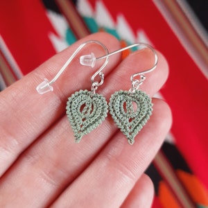 May include: A pair of silver earrings with green crocheted leaf-shaped charms. The earrings are hanging from a person's hand.
