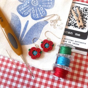 May include: Red crochet flower earrings with blue centres and green accents, alongside crochet hooks, spools of thread, and a printed card that says "scan me". The earrings are on a white cloth with blue floral patterns.