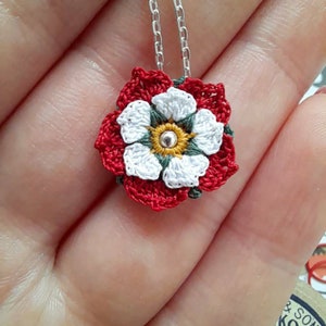 Op de afbeelding: Een gehaakte witte roos met een rode buitenlaag en een groen centrum, hangend aan een zilveren ketting. De roos is een hanger aan een ketting.