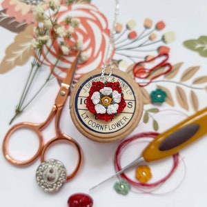 Op de afbeelding: Een ketting met een hanger in de vorm van een witte en rode geborduurde bloem met groene accenten. De hanger bevindt zich op een spoel garen met de tekst "J. Dewhurst & Sons Ltd. Lt. Cornflower".
