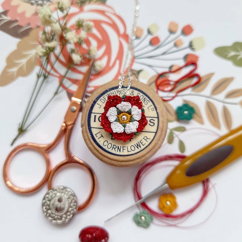 Op de afbeelding: Een ketting met een hanger in de vorm van een witte en rode geborduurde bloem met groene accenten. De hanger bevindt zich op een spoel garen met de tekst "J. Dewhurst & Sons Ltd. Lt. Cornflower".