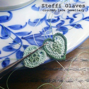 May include: Close-up of crochet heart-shaped lace jewelry in shades of green and white, with a crochet hook. The background features a blue and white floral patterned plate. Text reads "Steffi Glaves crochet lace jewellery."