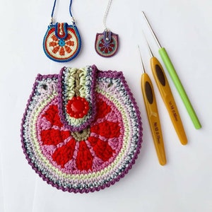 May include: A crocheted purse with a red, pink and green flower design. The purse has a button closure and is made with a variety of colours. Two crochet hooks are also pictured, one is labelled "No. 10 0.75mm" and the other is labelled "No. 14 0.50mm".