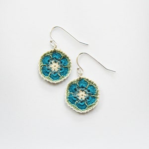 May include: A pair of crocheted flower earrings with a blue, green, and white color scheme. The earrings are attached to silver hooks.