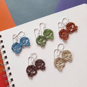May include: Set of six pairs of silver skull earrings. Each pair features a different color: blue, green, orange, brown, gold, and red.