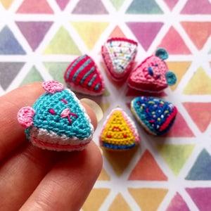 K&ouml;nnte beinhalten: Sechs geh&auml;kelte Mini-Kuchen in verschiedenen Farben und Designs. Die Kuchen sind dreieckig und haben verschiedene Bel&auml;ge, darunter Streusel, Streifen und ein Comic-Gesicht.