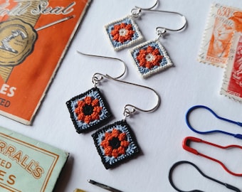 May include: A pair of crocheted earrings with a floral design. The earrings are made with orange, black, and white yarn. The earrings are hanging from silver hooks.
