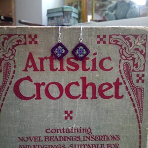 May include: A pair of silver earrings with purple and green crocheted flower designs. The earrings are displayed on a vintage book titled "Artistic Crochet" with the text "containing NOVEL BEADINGS, INSERTIONS AND EDGINGS SUITABLE FOR UNDERWEAR AND DRESS TRIMMINGS, EXQUISITE FLORAL DESIGNS IN IRISH".