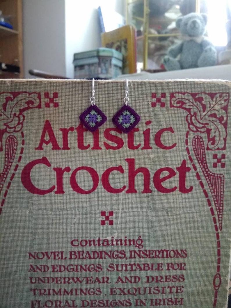 May include: A pair of silver earrings with purple and green crocheted flower designs. The earrings are displayed on a vintage book titled "Artistic Crochet" with the text "containing NOVEL BEADINGS, INSERTIONS AND EDGINGS SUITABLE FOR UNDERWEAR AND DRESS TRIMMINGS, EXQUISITE FLORAL DESIGNS IN IRISH".