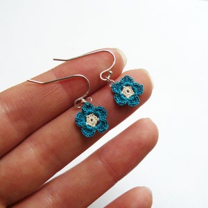 May include: A pair of handmade crochet flower earrings. Each earring features a teal blue flower with a cream-colored center. The earrings have silver-colored ear wires. The earrings are small and delicate, perfect for everyday wear.