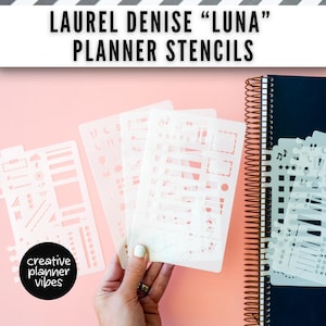 May include: A set of translucent plastic Laurel Denise Luna planner stencils featuring various cut-out shapes including boxes, lines, stars, and musical notes, held in a hand next to a black spiral-bound planner on a light pink background.
