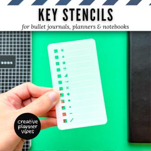 Puede incluir: Un esténcil de llave de plástico blanco con varias formas y símbolos, incluyendo estrellas, cuadrados y flechas. El texto "KEY STENCILS" y "for bullet journals, planners & notebooks" están en la parte superior. El esténcil se sostiene en una mano sobre un fondo verde.
