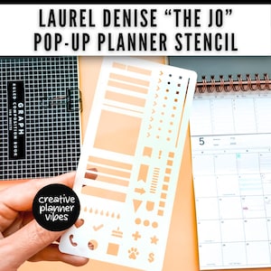 以下が含まれることがあります： 長方形、矢印、星など、さまざまな形やアイコンが描かれた白いプラスチック製のプランナーステンシルが、方眼ノートとカレンダーの前に置かれています。「LAUREL DENISE 'THE JO' POP-UP PLANNER STENCIL」というテキストが上部にあります。