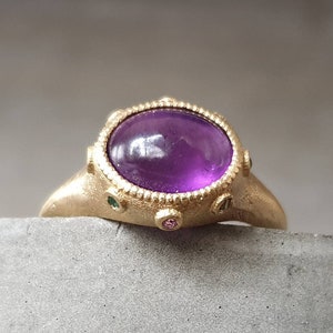K&ouml;nnte beinhalten: Ein goldener Ring mit einem gro&szlig;en violetten Amethyst-Edelstein, umgeben von kleineren gr&uuml;nen und rosa Edelsteinen.
