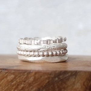 May include: A stack of four silver rings with distinct textures. The top ring has a geometric notched edge, the second has a rough organic surface, the third features a beaded design, and the bottom ring has a wide, hammered, matte finish.