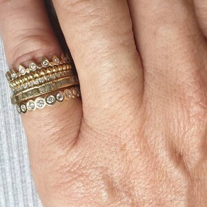 以下が含まれることがあります： 小さなダイヤモンドがついた金の指輪を5つ重ねてつけた手。指輪はそれぞれデザインが異なり、小さなダイヤモンドが並んだ指輪、小さな三角形が並んだ指輪、小さな円が並んだ指輪などがあります。