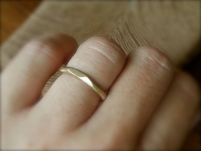 May include: A gold band ring with a hammered texture. The ring is worn on a finger.