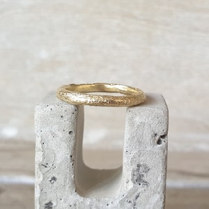 May include: A gold ring with a textured surface, resting on a gray concrete block. The ring has a band with a bark-like texture, and the gold color is bright and shiny. The ring is a simple band style, suitable for everyday wear.