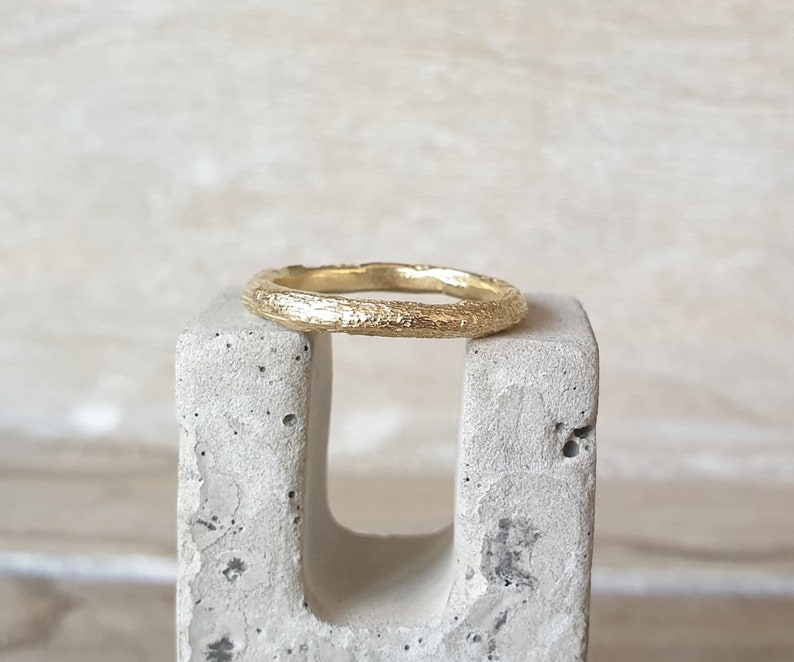 May include: A gold ring with a textured surface, resting on a gray concrete block. The ring has a band with a bark-like texture, and the gold color is bright and shiny. The ring is a simple band style, suitable for everyday wear.