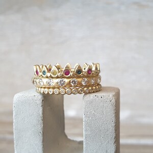 Peut inclure: Trois bagues dor&eacute;es empil&eacute;es sur un bloc de b&eacute;ton gris. La bague sup&eacute;rieure a un motif de couronne avec des pierres pr&eacute;cieuses rouges, vertes et jaunes altern&eacute;es. La bague du milieu comporte des pierres pr&eacute;cieuses claires et la bague inf&eacute;rieure a de petites pierres pr&eacute;cieuses claires.