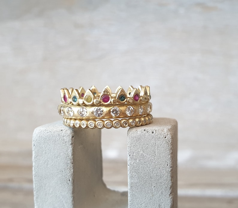 Peut inclure: Trois bagues dor&eacute;es empil&eacute;es sur un bloc de b&eacute;ton gris. La bague sup&eacute;rieure a un motif de couronne avec des pierres pr&eacute;cieuses rouges, vertes et jaunes altern&eacute;es. La bague du milieu comporte des pierres pr&eacute;cieuses claires et la bague inf&eacute;rieure a de petites pierres pr&eacute;cieuses claires.