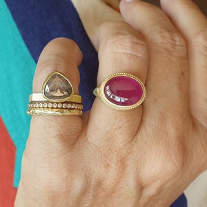 May include: A hand wearing several gold rings. One ring features a large, oval, ruby-colored stone set in a gold band. Another ring has a triangular, smoky-colored stone. Other rings are gold bands.