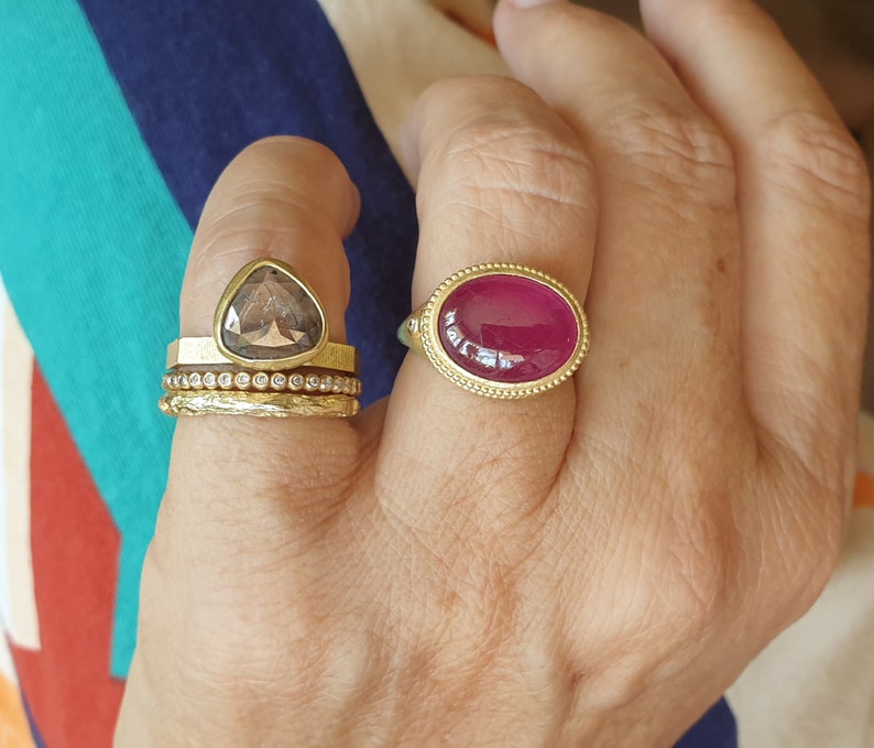 May include: A hand wearing several gold rings. One ring features a large, oval, ruby-colored stone set in a gold band. Another ring has a triangular, smoky-colored stone. Other rings are gold bands.