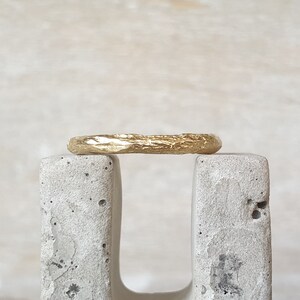 May include: A gold ring with a textured, organic design. The ring is displayed on a gray, concrete block against a neutral background. The ring's surface has a rough, uneven texture, giving it a unique, handcrafted appearance.