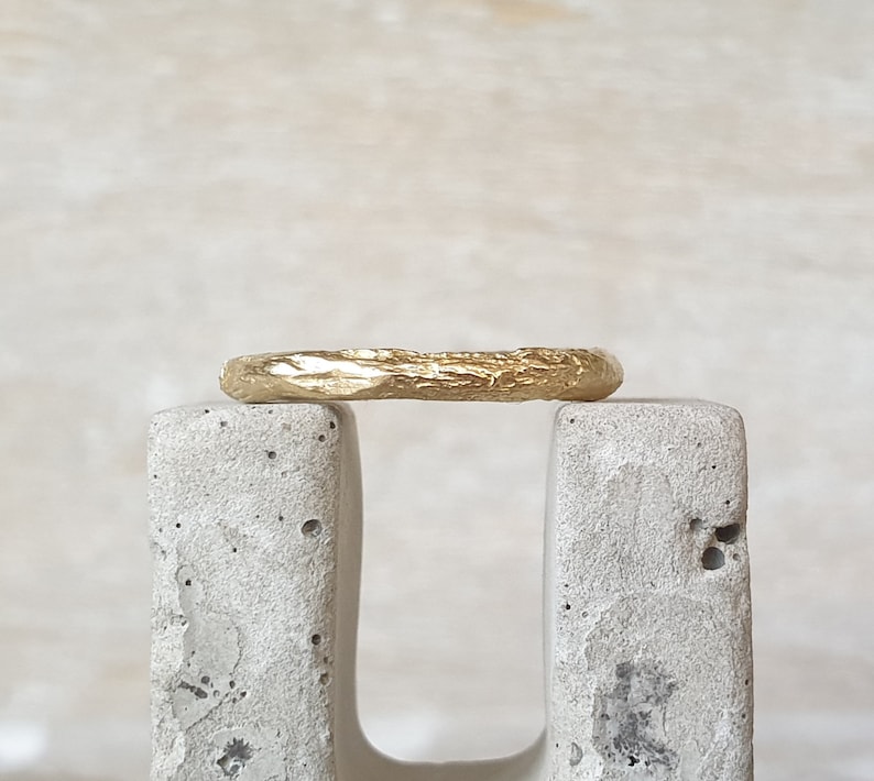 May include: A gold ring with a textured, organic design. The ring is displayed on a gray, concrete block against a neutral background. The ring's surface has a rough, uneven texture, giving it a unique, handcrafted appearance.