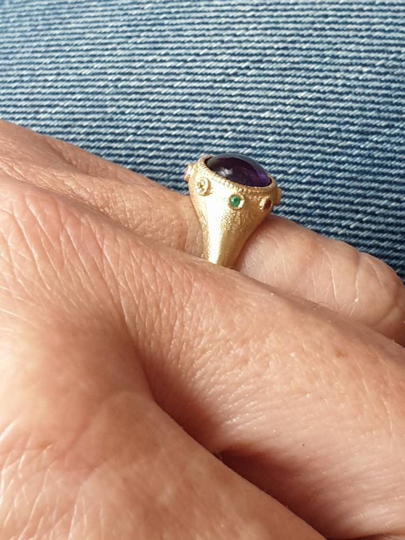 K&ouml;nnte beinhalten: Ein goldener Ring mit einem gro&szlig;en violetten Edelstein, der von kleineren gr&uuml;nen und roten Edelsteinen umgeben ist.