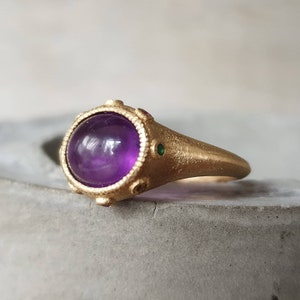 K&ouml;nnte beinhalten: Ein goldener Ring mit einem gro&szlig;en, runden, violetten Amethyst-Edelstein, der in einer Zargenfassung sitzt. Der Ring hat eine strukturierte Band und ist mit kleinen gr&uuml;nen und roten Edelsteinen verziert.
