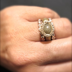 May include: A gold ring with a large, oval-shaped, grey gemstone surrounded by smaller diamonds. The ring is worn on a person's finger.