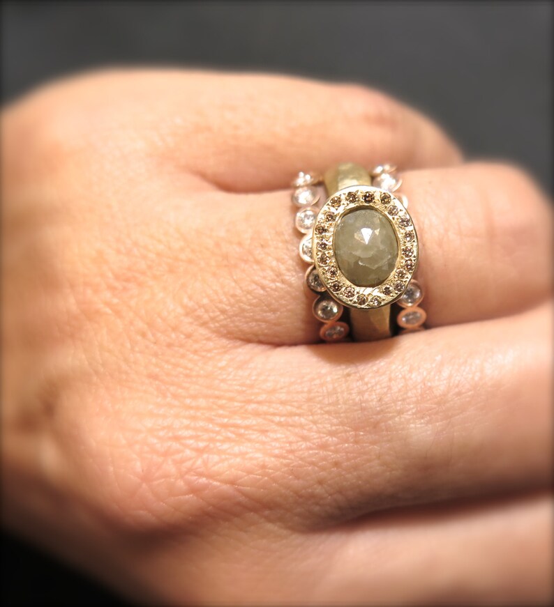 May include: A gold ring with a large, oval-shaped, grey gemstone surrounded by smaller diamonds. The ring is worn on a person's finger.