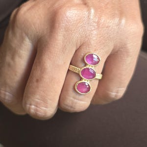 May include: A gold ring featuring a textured, beaded band and three vertically aligned, oval-shaped, faceted pink gemstones. The stones are set in gold bezels, with the middle stone positioned slightly higher than the top and bottom stones.