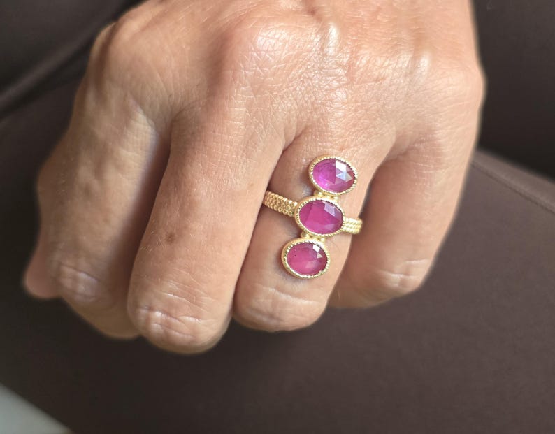May include: A gold ring featuring a textured, beaded band and three vertically aligned, oval-shaped, faceted pink gemstones. The stones are set in gold bezels, with the middle stone positioned slightly higher than the top and bottom stones.