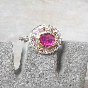 May include: A silver ring with a large, oval, magenta gemstone at the center, surrounded by smaller pink and orange gemstones. The ring has a gold-colored setting around the central stone and is set on a gray surface.