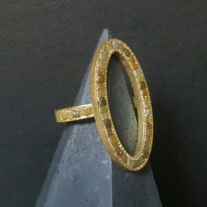 May include: A gold ring with a large oval-shaped setting. The setting is filled with small, irregular-shaped diamonds. The ring is on a gray, textured surface.