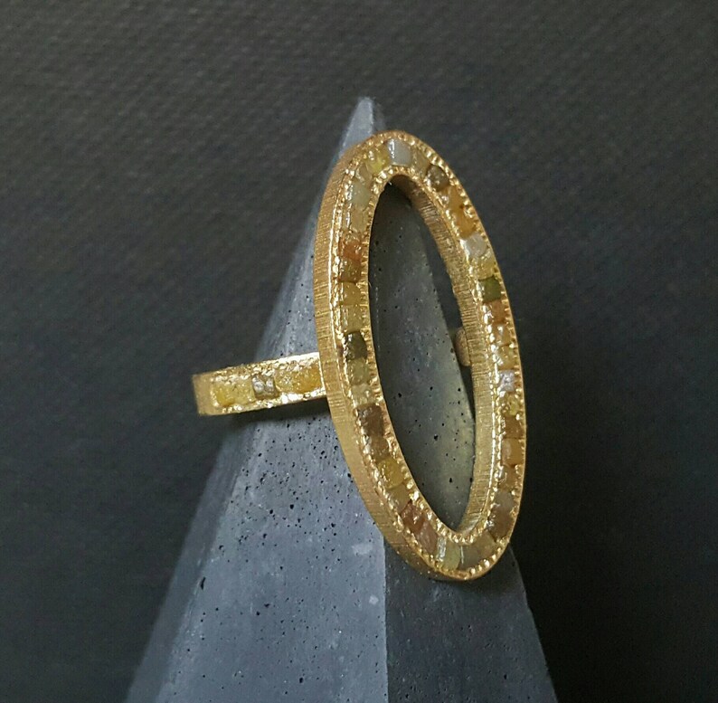 May include: A gold ring with a large oval-shaped setting. The setting is filled with small, irregular-shaped diamonds. The ring is on a gray, textured surface.