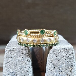 Peut inclure: Trois bagues en or empil&eacute;es. La bague sup&eacute;rieure est orn&eacute;e de pierres vertes, la bague du milieu de pierres blanches et la bague inf&eacute;rieure de petites pierres vertes. Les bagues sont pos&eacute;es sur une surface grise.
