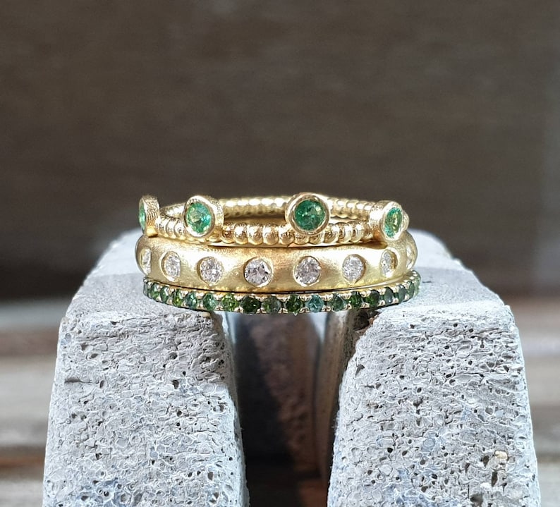 Peut inclure: Trois bagues en or empil&eacute;es. La bague sup&eacute;rieure est orn&eacute;e de pierres vertes, la bague du milieu de pierres blanches et la bague inf&eacute;rieure de petites pierres vertes. Les bagues sont pos&eacute;es sur une surface grise.