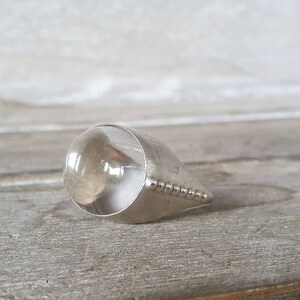 May include: A silver-toned ring with a large, clear, rounded stone. The ring has a teardrop shape, with a band of small, raised dots along the edge. The stone appears to be transparent, with a slight amber hue.