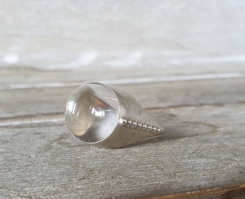 May include: A silver-toned ring with a large, clear, rounded stone. The ring has a teardrop shape, with a band of small, raised dots along the edge. The stone appears to be transparent, with a slight amber hue.