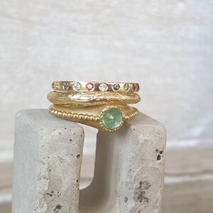 May include: A stack of gold rings with colorful gemstones. The top ring has a row of small, evenly spaced gems in various colors. The middle ring has a textured surface. The bottom ring features a large, round, green gemstone.