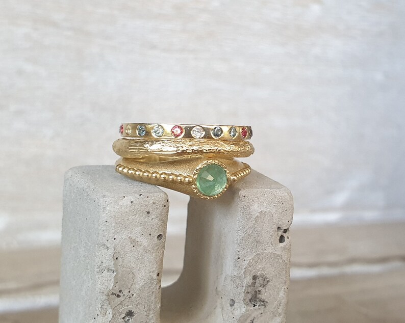 May include: A stack of gold rings with colorful gemstones. The top ring has a row of small, evenly spaced gems in various colors. The middle ring has a textured surface. The bottom ring features a large, round, green gemstone.