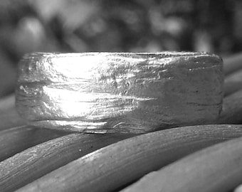 May include: Close-up of a silver ring with a textured, bark-like surface. The ring is thick and has a wide band. The ring is resting on a surface of thin, wooden dowels.