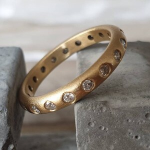 Peut inclure: Une bague en or avec une finition mate, sertie de nombreux petits pierres pr&eacute;cieuses transparentes. La bague est pr&eacute;sent&eacute;e sur une surface grise, mettant en valeur son design circulaire et l'&eacute;clat des pierres. La bande de la bague mesure environ 0,6 cm de large.