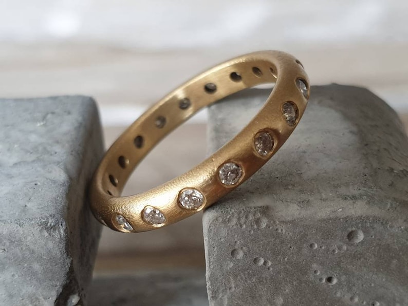 Peut inclure: Une bague en or avec une finition mate, sertie de nombreux petits pierres pr&eacute;cieuses transparentes. La bague est pr&eacute;sent&eacute;e sur une surface grise, mettant en valeur son design circulaire et l'&eacute;clat des pierres. La bande de la bague mesure environ 0,6 cm de large.
